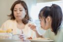 Seorang ibu sedang mendampingi buah hatinya menikmati makanan bergizi dalam suasana yang hangat dan penuh kasih sayang di rumah.