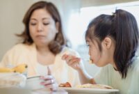 Seorang ibu sedang mendampingi buah hatinya menikmati makanan bergizi dalam suasana yang hangat dan penuh kasih sayang di rumah.