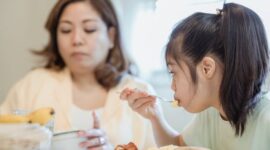 Seorang ibu sedang mendampingi buah hatinya menikmati makanan bergizi dalam suasana yang hangat dan penuh kasih sayang di rumah.